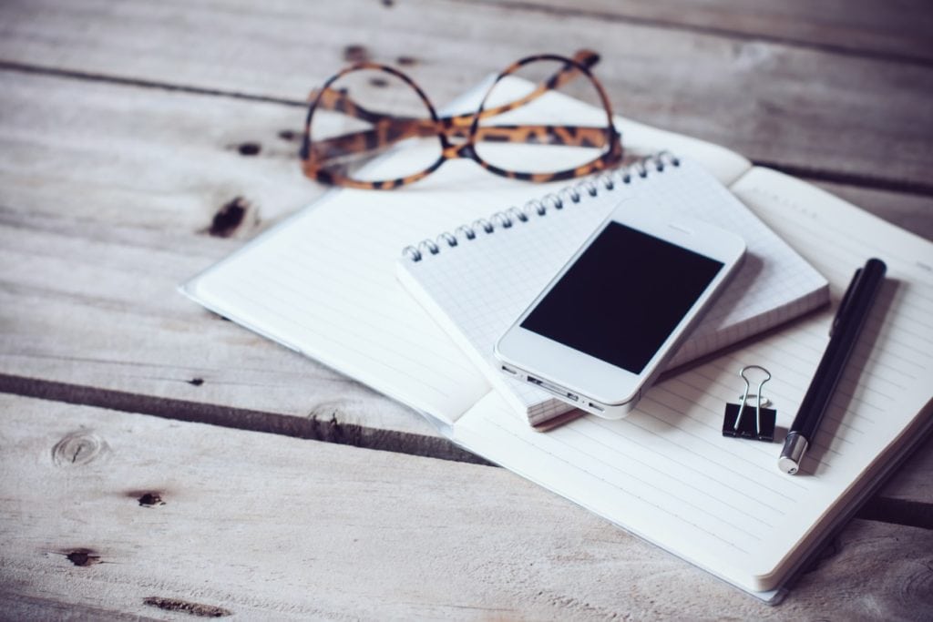 Hipster home office tabletop: papers and notebooks, reading glasses, smart phone, pen on an old wooden board background. Vintage lifestyle.